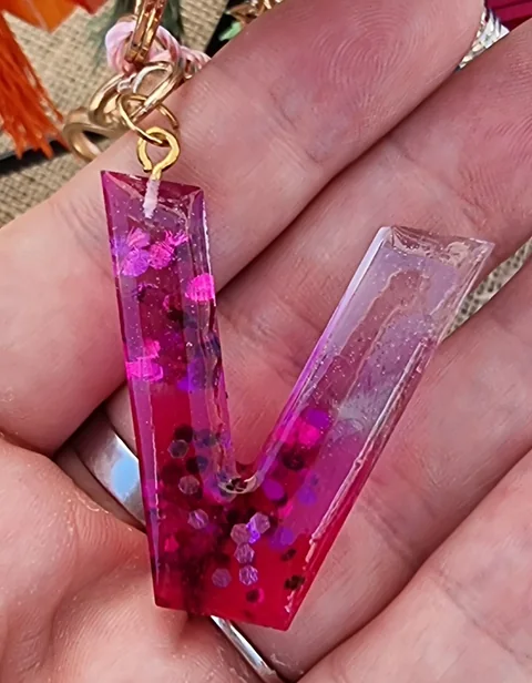 Lettera V in resina rosa trasparente con inclusioni decorative, tenuta su una mano